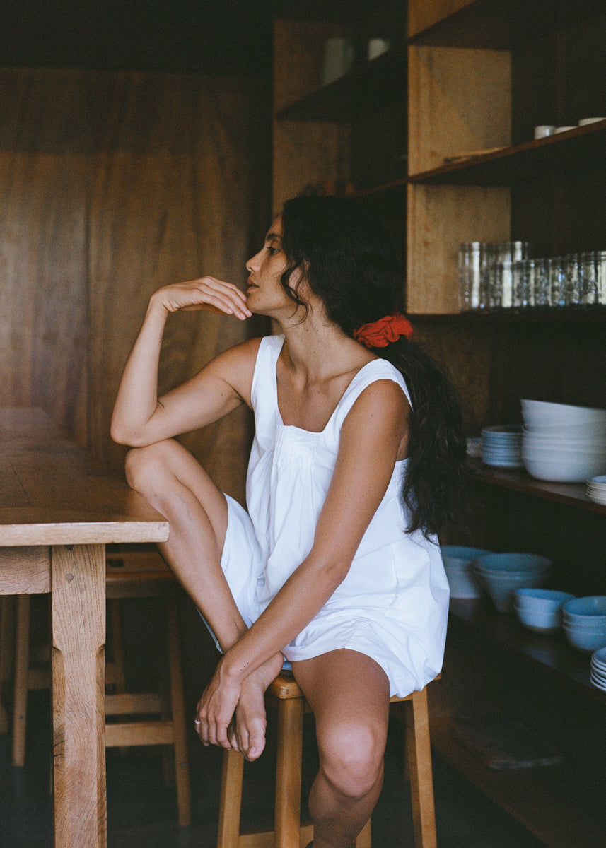 Woman in a white dress sitting on a wooden stool in a rustic kitchen.