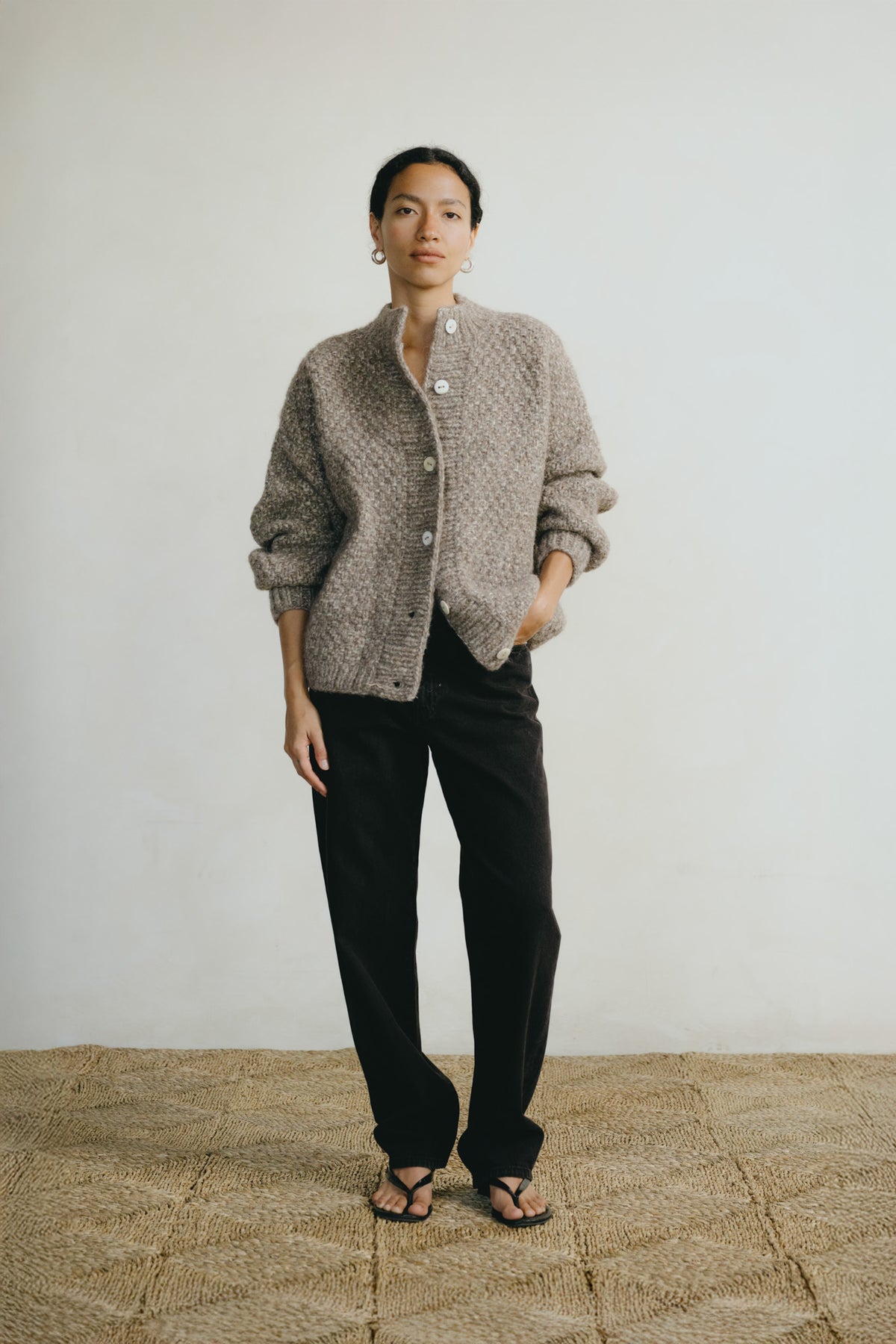 A model stands against a plain off-white wall wearing a textured, light taupe-gray knit cardigan with white buttons, black trousers, and black sandals.