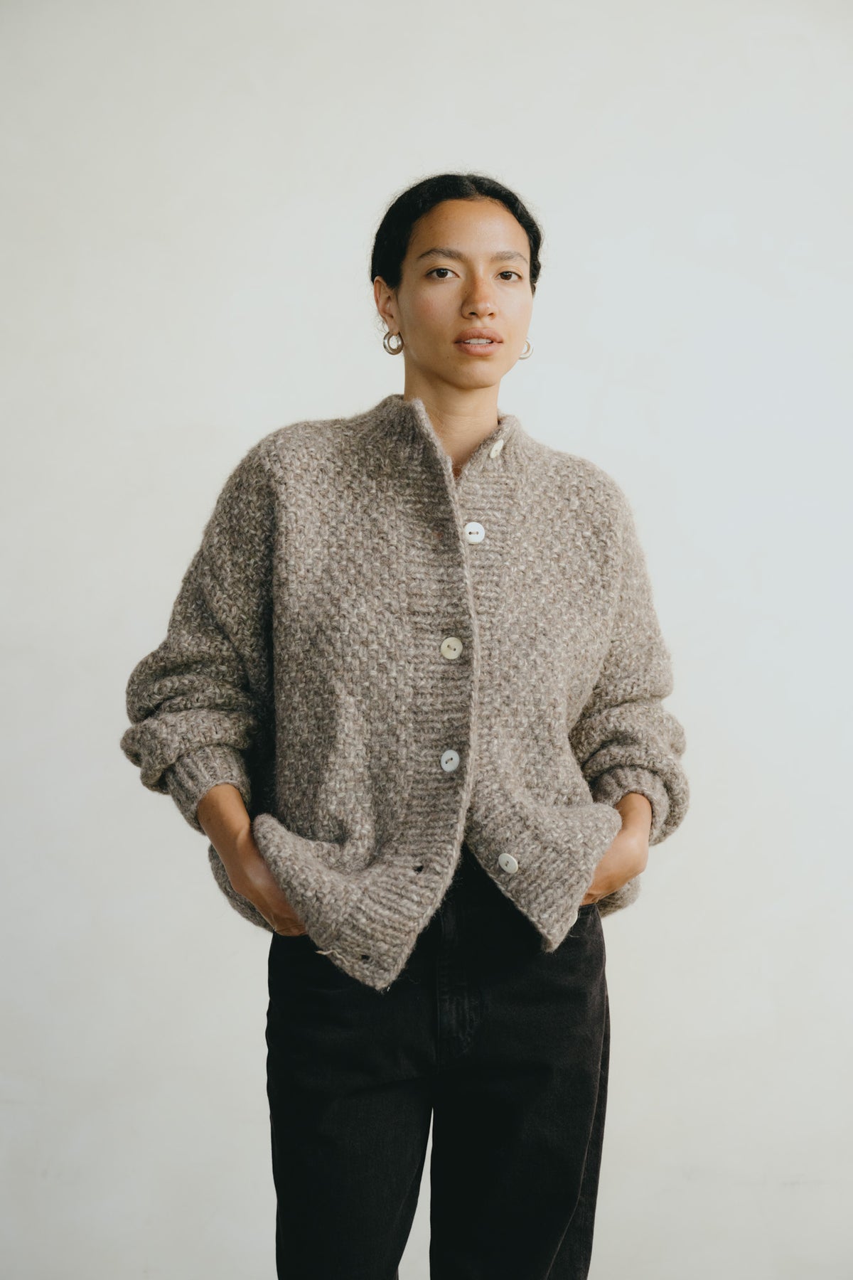 A woman wearing a chunky knit cardigan sweater in a taupe-gray color with white buttons, paired with black pants, standing against a plain light gray background.