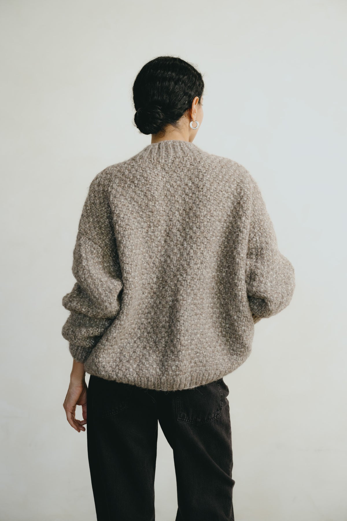 A person shown from the back wearing a chunky knit sweater in a warm taupe-beige color, paired with black pants, against a plain off-white background.