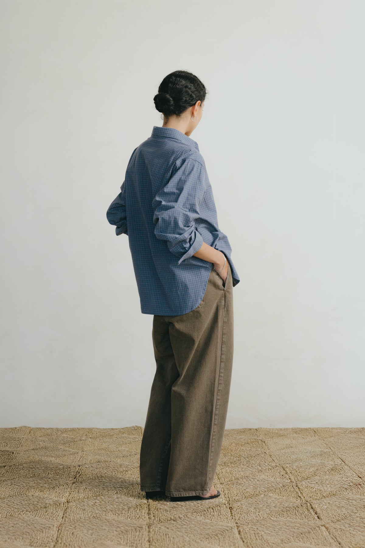 A person standing with their back to the camera, wearing a loose blue button-up shirt and wide-leg brown pants, set against a plain off-white wall and a woven straw floor.