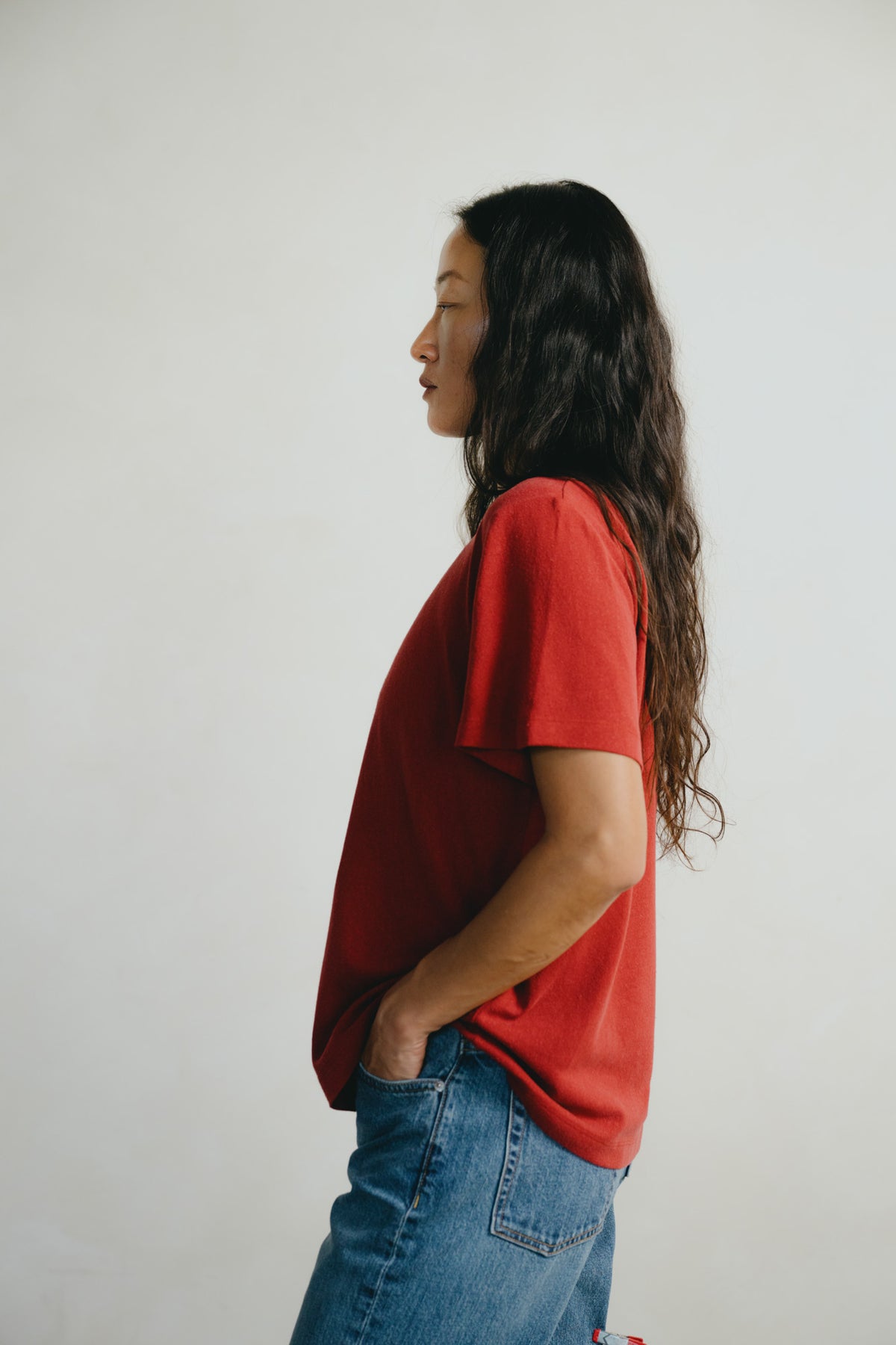 A woman with long dark hair is shown in profile wearing a loose red short-sleeve t-shirt and blue jeans against a plain light gray background.