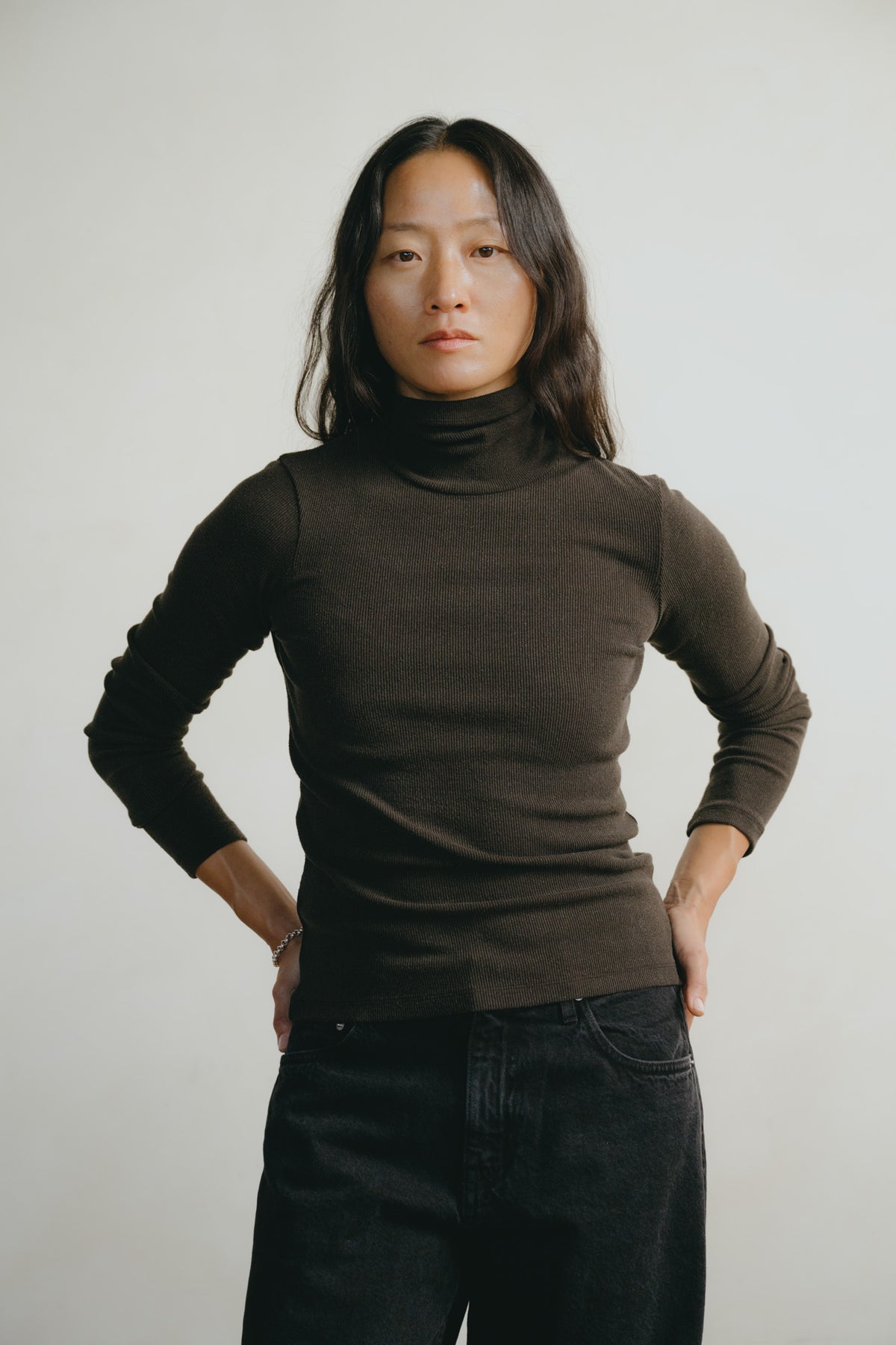 A woman with shoulder-length dark hair wearing a dark brown ribbed turtleneck sweater and dark jeans, standing against a light gray studio background with her hands on her hips.
