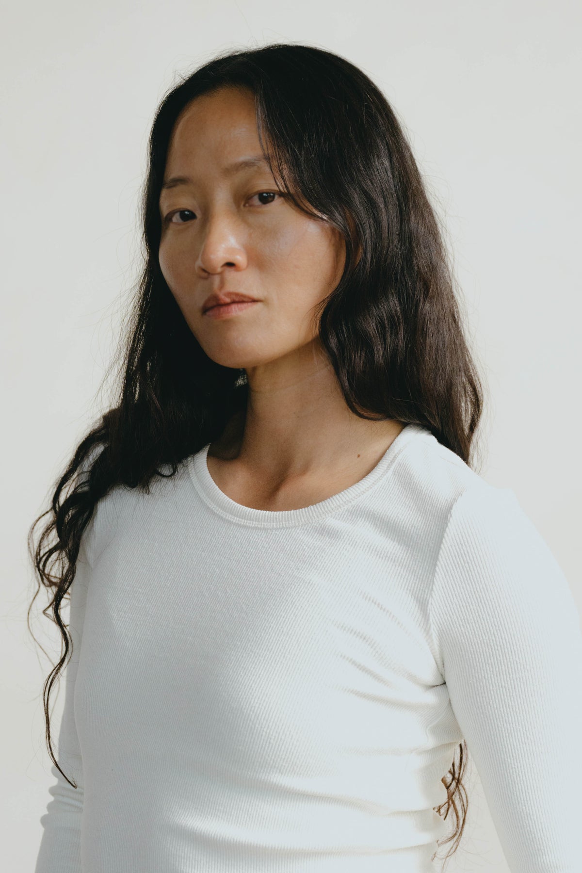 A woman with long dark hair wearing a white long-sleeve crewneck top, photographed against a plain light background.