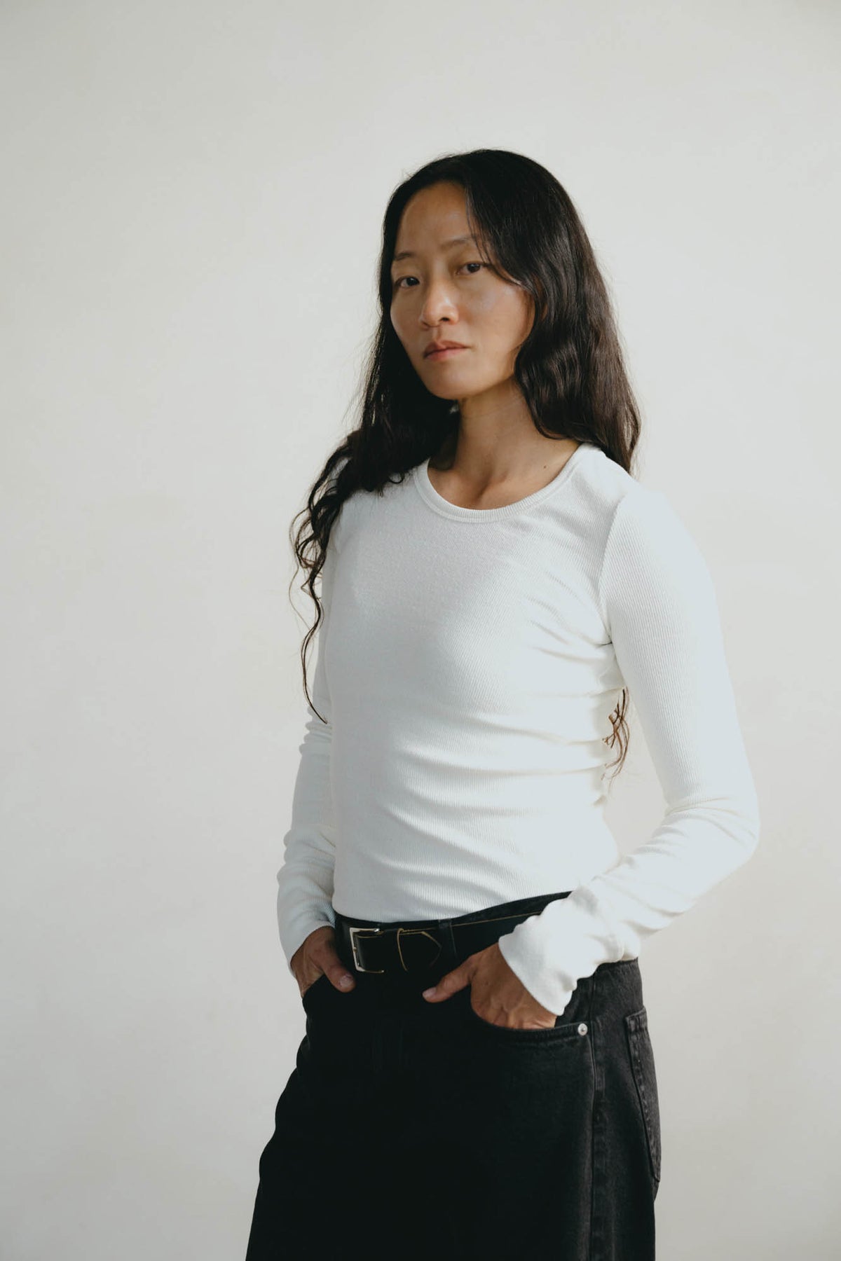 A woman with long dark hair wearing a fitted white long-sleeve crewneck top and black jeans with a black belt, standing against a plain off-white background.