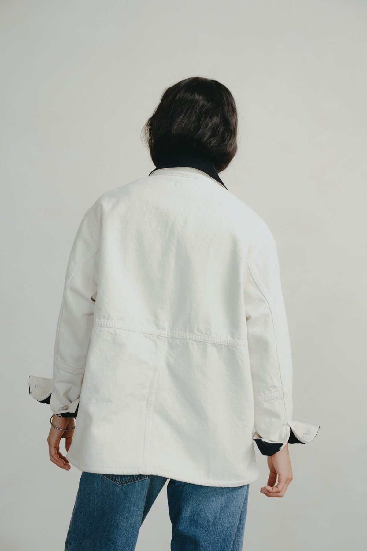 A person shown from behind wearing an oversized white jacket with a black collar and black cuffs, paired with blue jeans, against a plain light gray background.