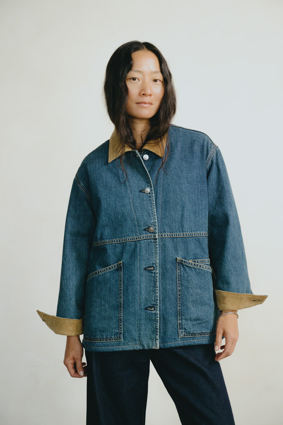 A model wearing a blue denim jacket with tan contrast collar, cuffs, and front patch pockets, paired with dark pants, against a plain light gray studio background.