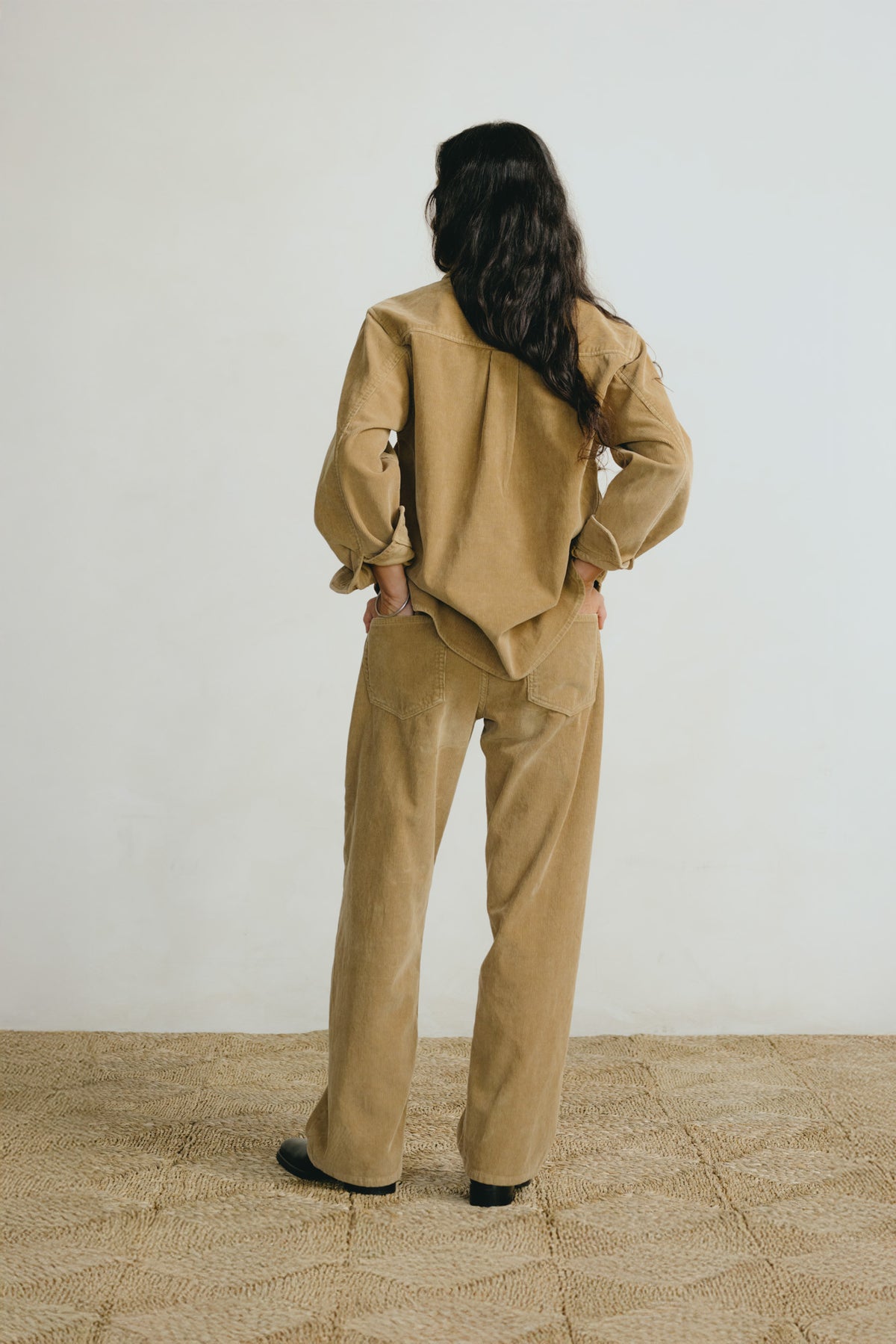A woman stands with her back to the camera wearing a matching tan corduroy jacket and wide-leg trousers set, paired with black shoes, in a minimalist studio setting.