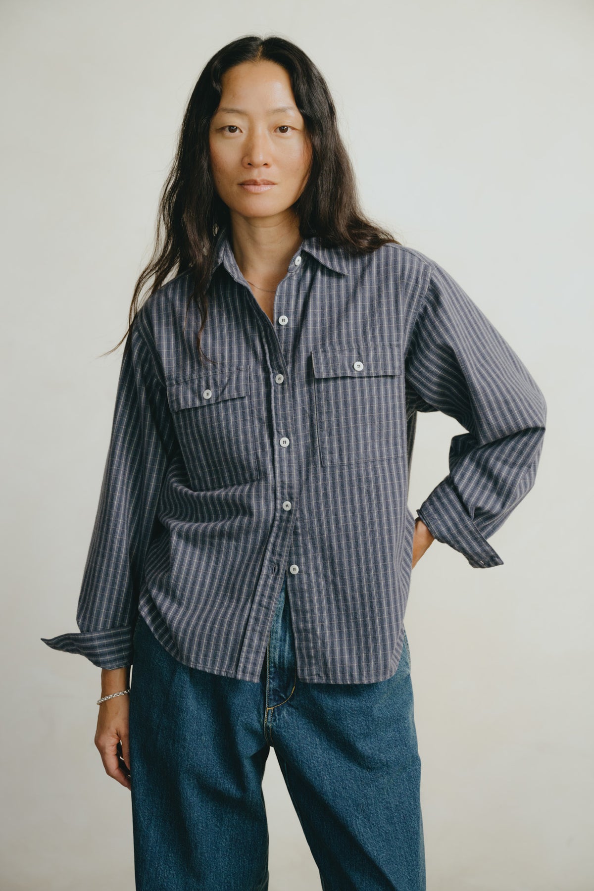 A woman modeling a dark gray and navy blue vertically striped button-up shirt with two chest pockets, paired with blue jeans, against a plain light gray background.