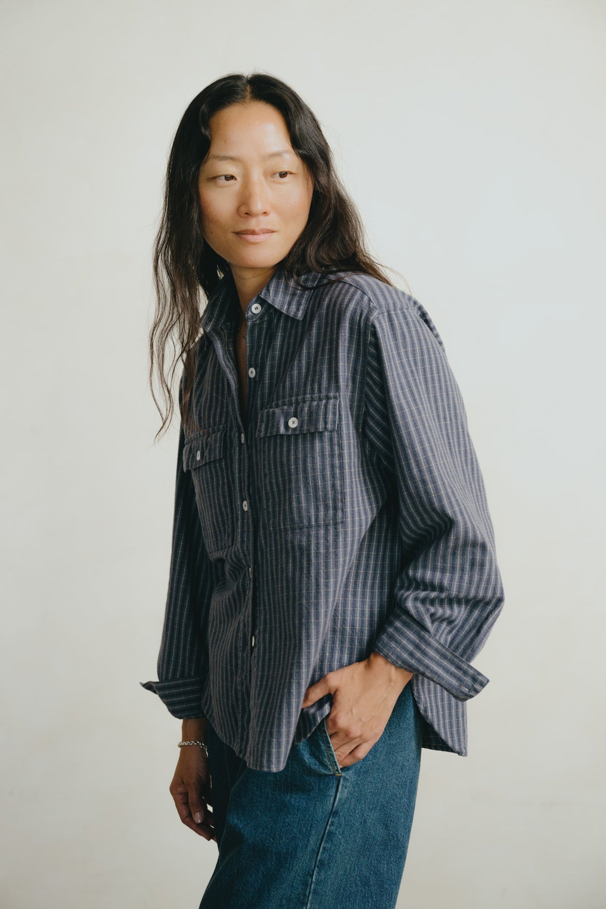 A woman wearing a dark navy and gray vertically striped button-down shirt with blue jeans, standing against a plain light gray background.