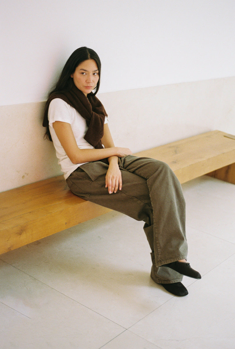 A woman sits on a light wood bench against a white wall, wearing a white short-sleeve top, brown pants, and black socks in a minimalist indoor setting.