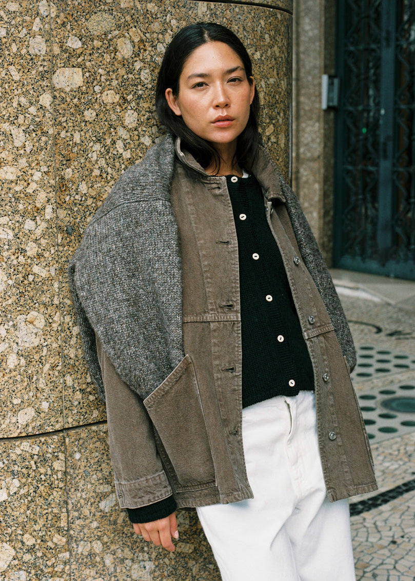 A model stands outdoors against a textured stone wall, wearing a layered jacket with a gray wool-like shoulder panel and a brown corduroy body, layered over a black buttoned top and white pants.