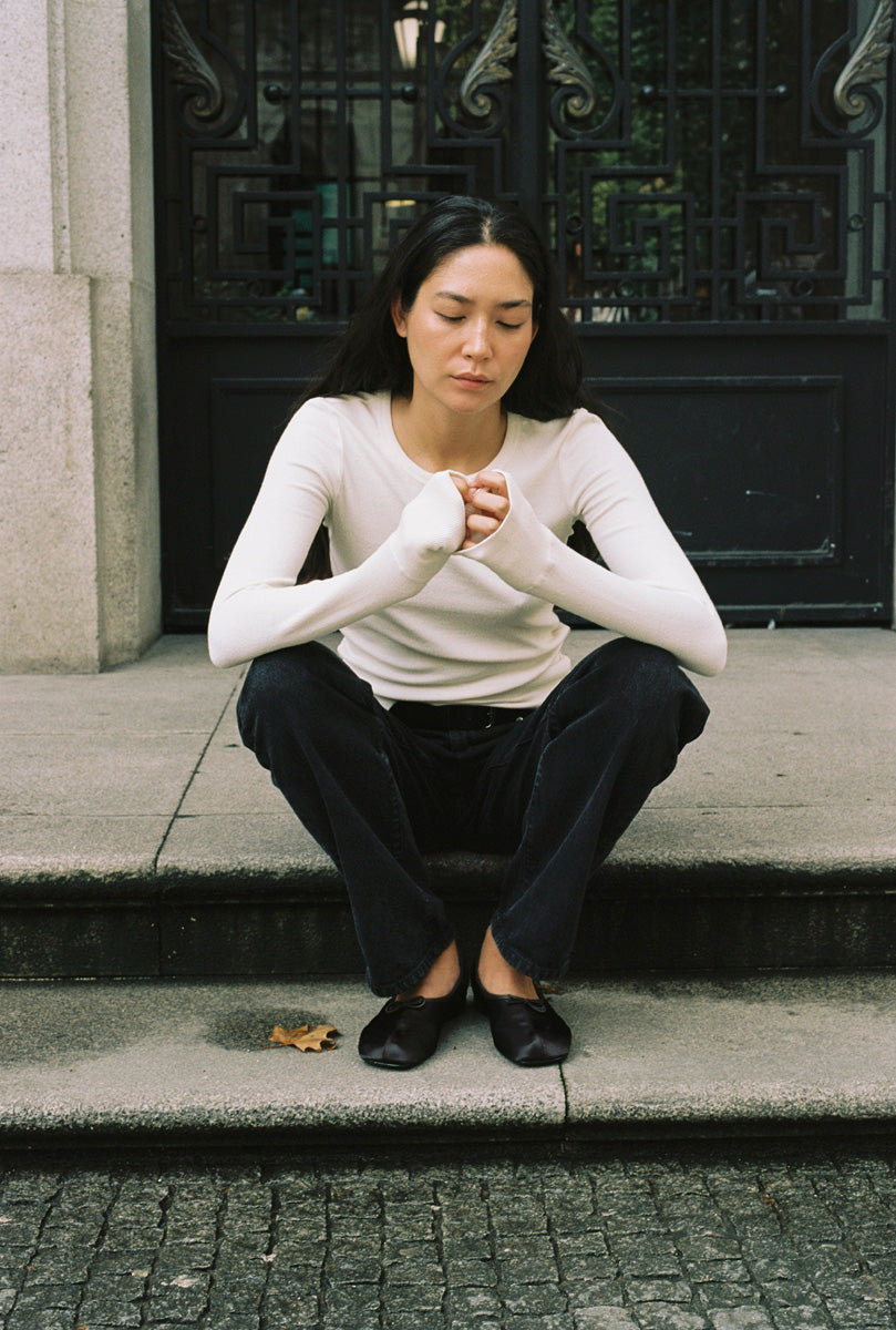 A woman sitting in a doorway with a long sleeve tight white shirt and black jeans.