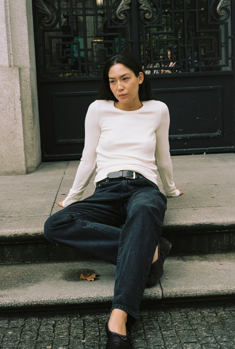 A woman sitting in a doorway with a long sleeve tight white shirt and black jeans.