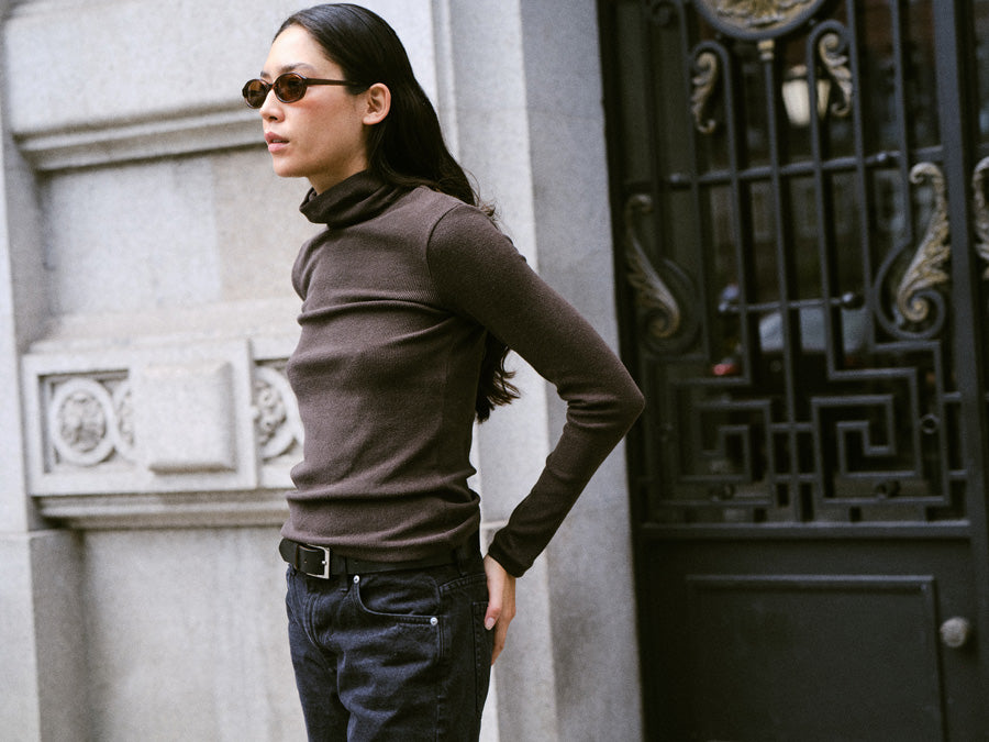 A woman standing on the street with sunglasses and wearing a long-sleeved dark brown turtleneck and black jeans.