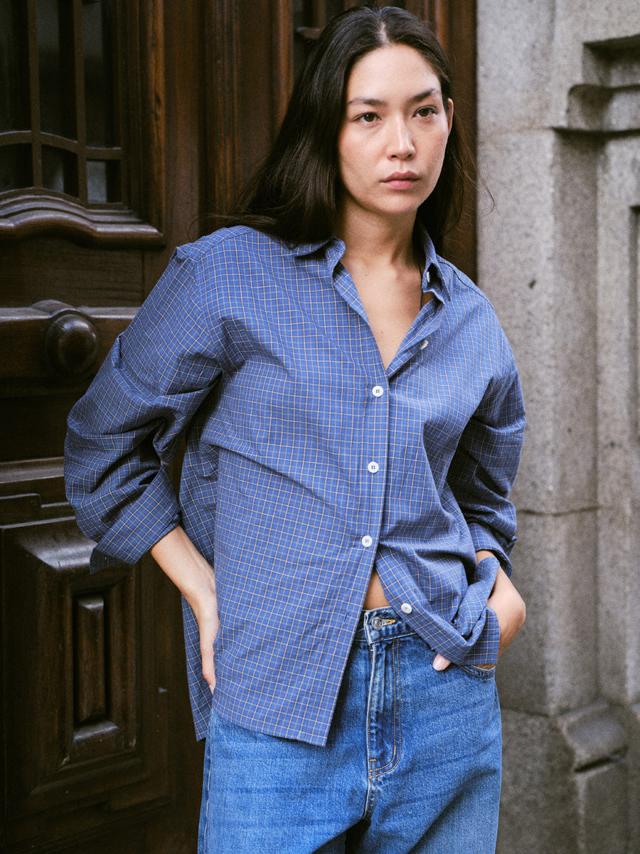 A woman wearing an oversized blue and red checkered button-down shirt with blue jeans, standing in front of a dark wooden door and stone wall.