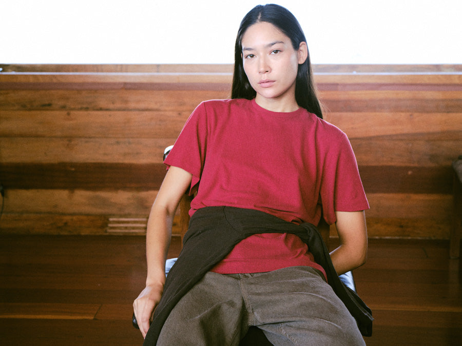 A woman sitting in a red tshirt against a wood wall, with brown pants.