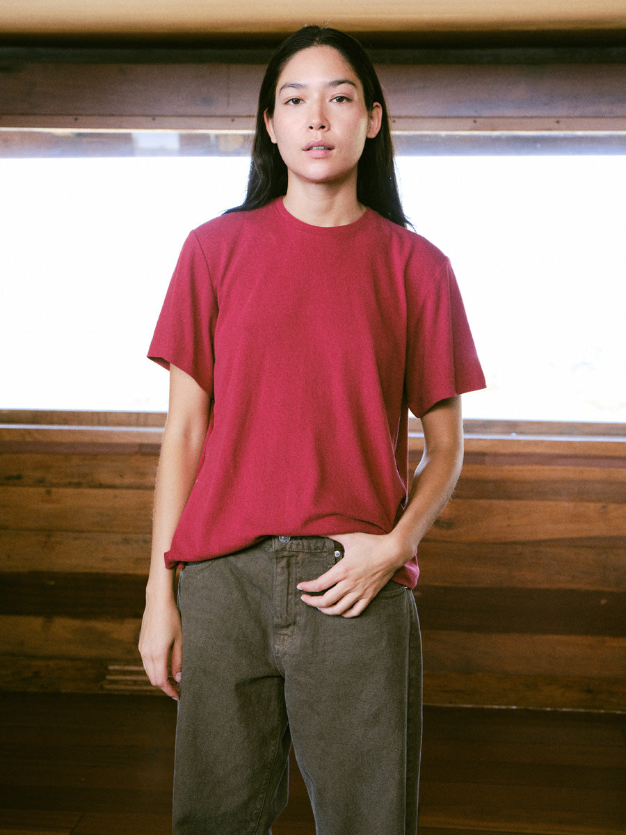 A woman standing in a red tshirt against a wood wall, with brown pants.