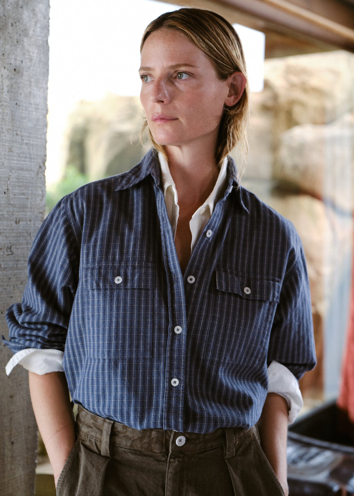 A woman wearing a blue and white vertically striped button-up shirt over a white undershirt, paired with brown trousers, standing in a rustic stone-walled environment.