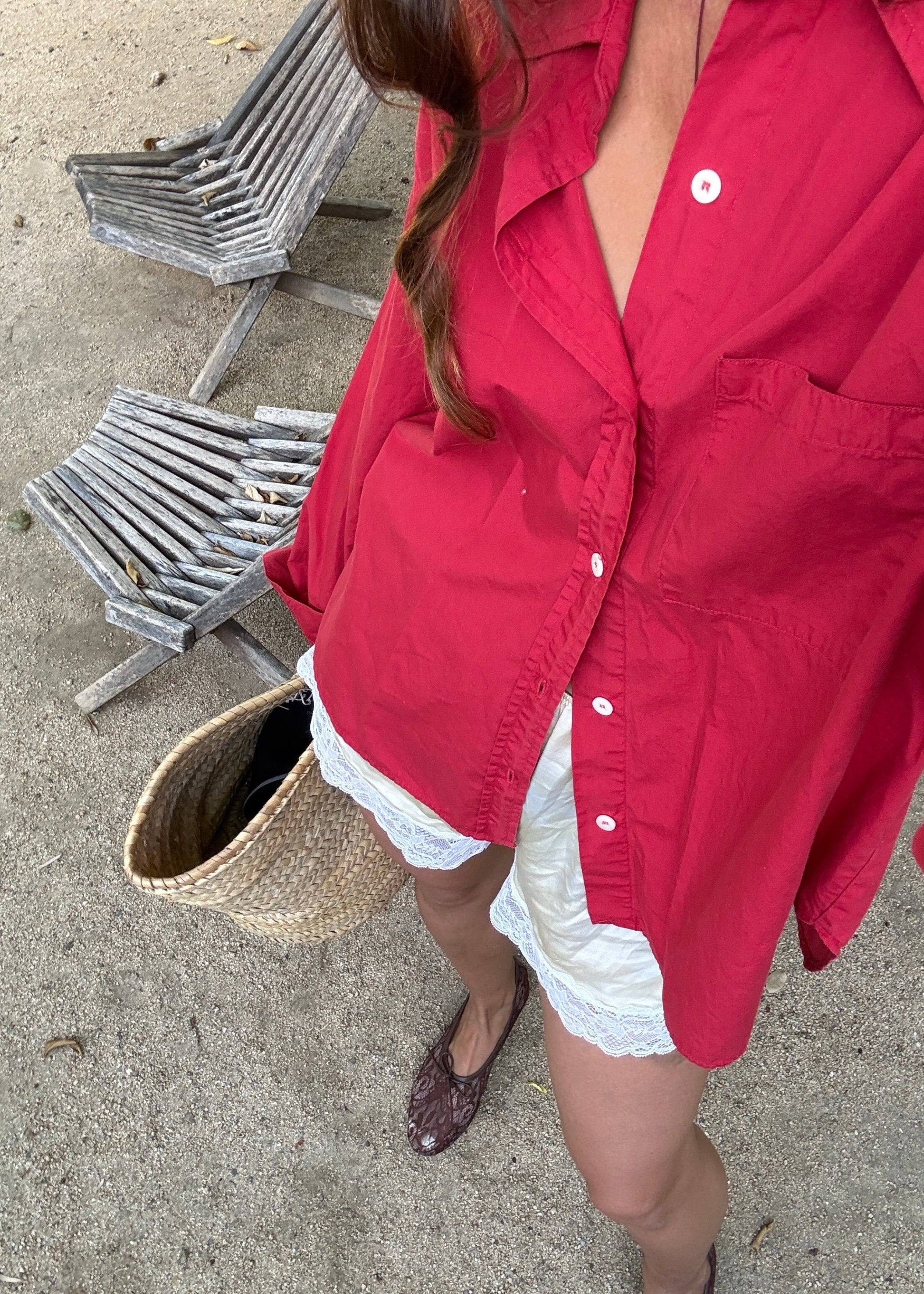 A person wearing an oversized red button-up shirt and white shorts, holding a woven straw bag outdoors on a concrete surface with wooden chairs in the background.