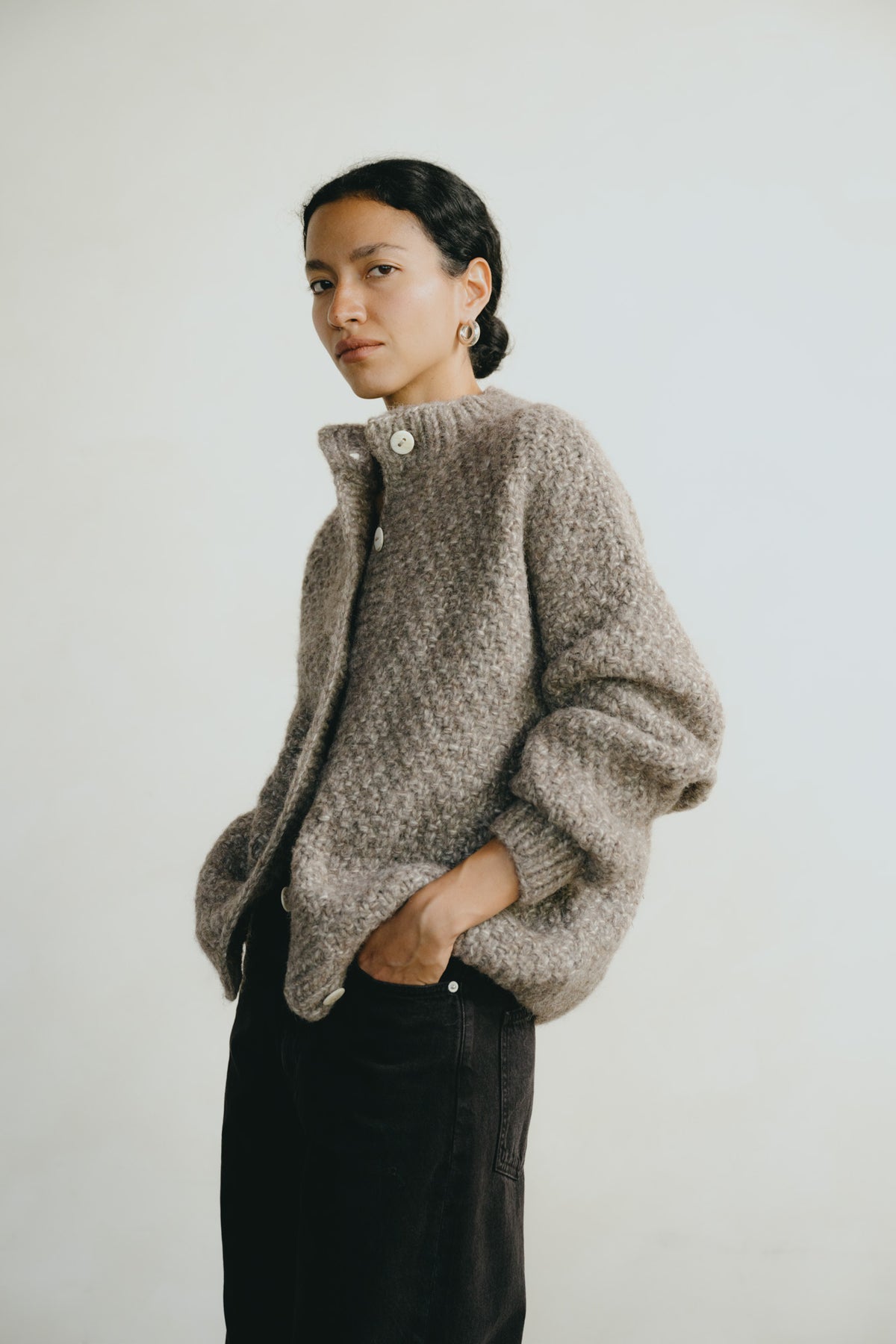 A woman wearing a textured, chunky knit cardigan in a taupe-gray color, paired with black pants, standing against a plain light gray background.