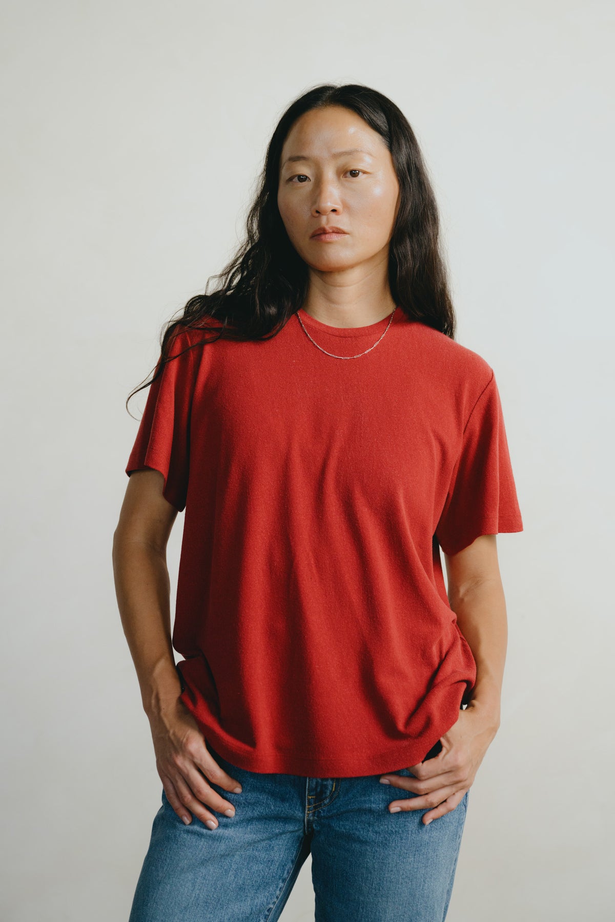 A woman with long dark hair wearing a red short-sleeve crewneck t-shirt and blue jeans, standing against a plain light gray background.