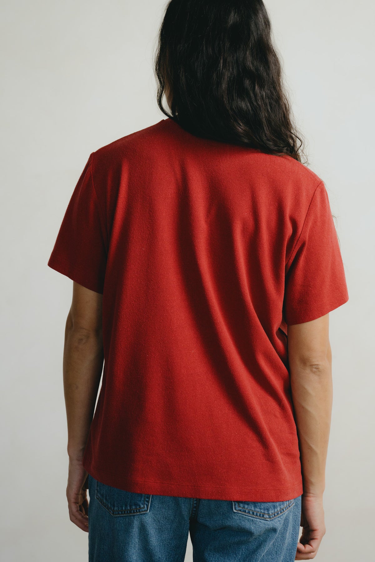 A person viewed from behind wearing a solid red short-sleeve t-shirt and blue jeans against a plain light gray background.