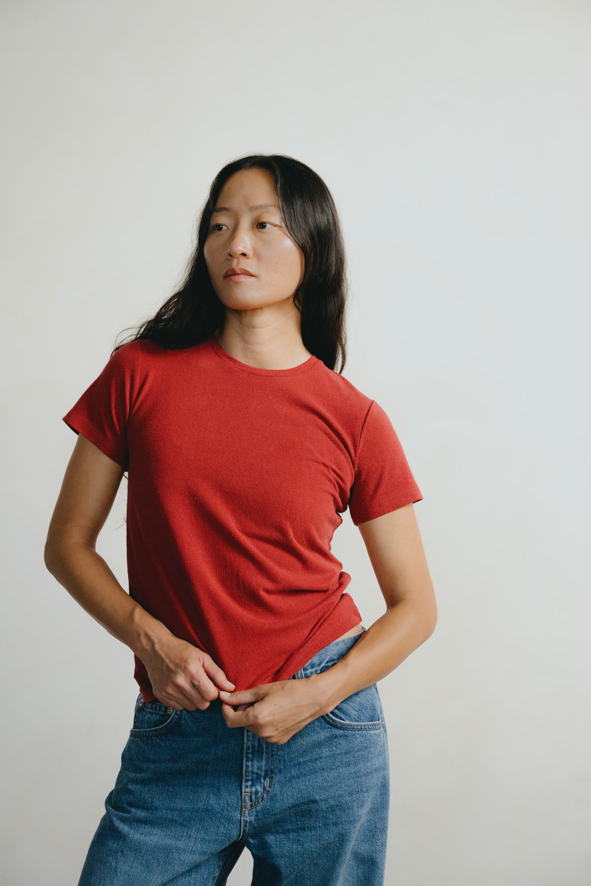 A woman wearing a red short-sleeve crewneck t-shirt and blue jeans, standing against a plain light gray background.