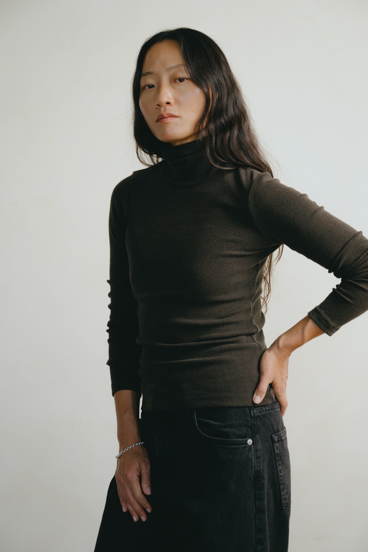 A woman modeling a dark brown ribbed turtleneck sweater, paired with dark jeans, against a plain light gray background.