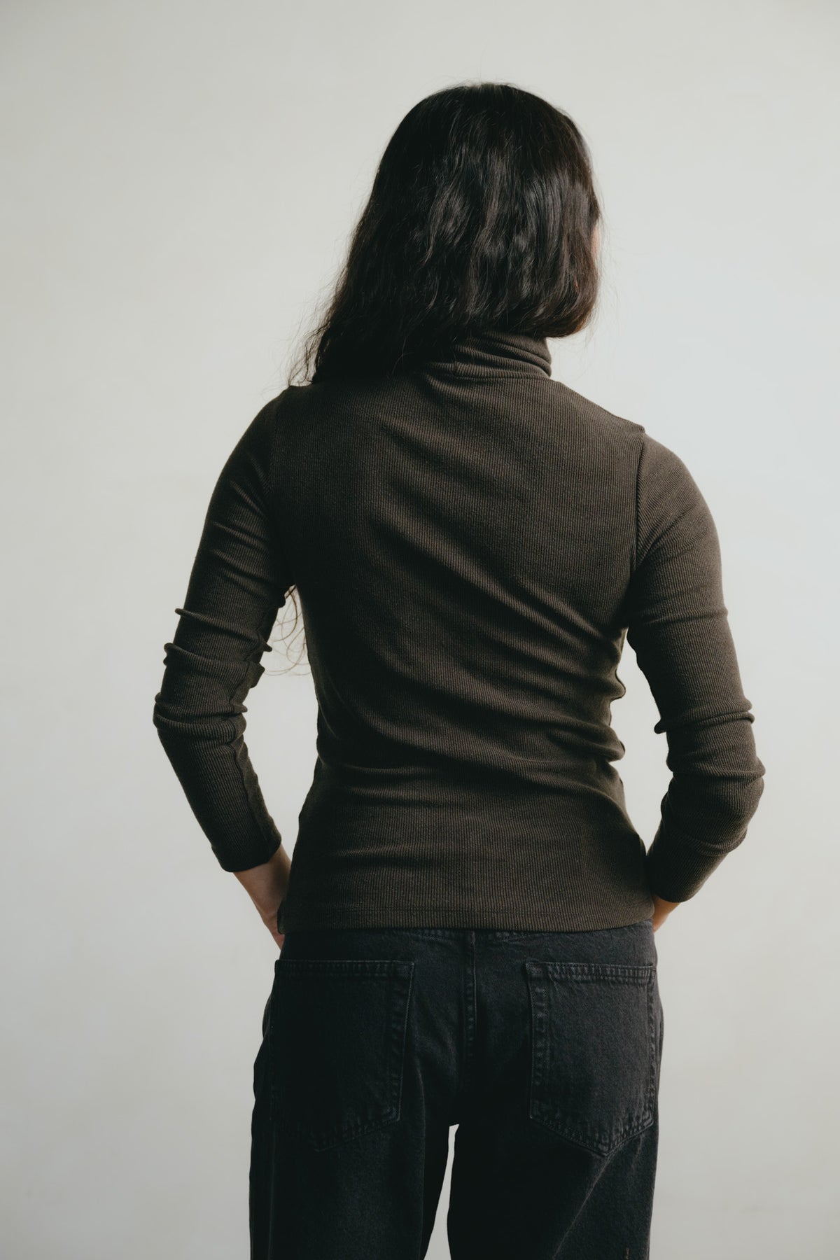 A person shown from the back wearing a fitted dark brown long-sleeve turtleneck top and dark jeans against a plain light gray background.