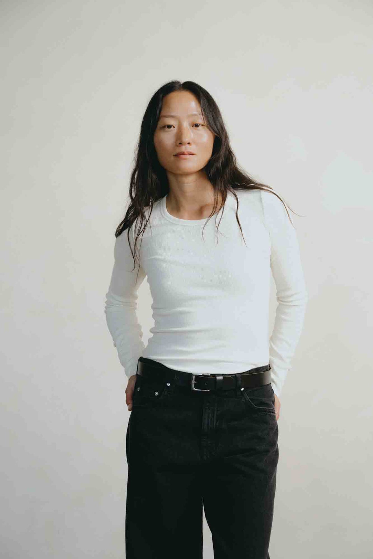 A woman with long dark hair stands against a plain light background wearing a fitted white long-sleeve top and black jeans with a black belt.