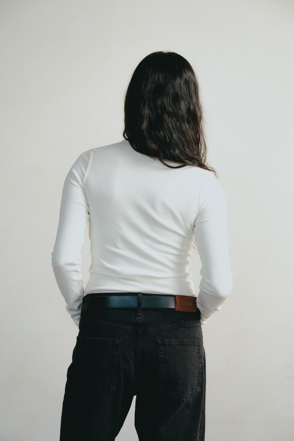 A person viewed from behind wearing a fitted white long-sleeve top and dark jeans with a dark belt, standing against a plain light background.