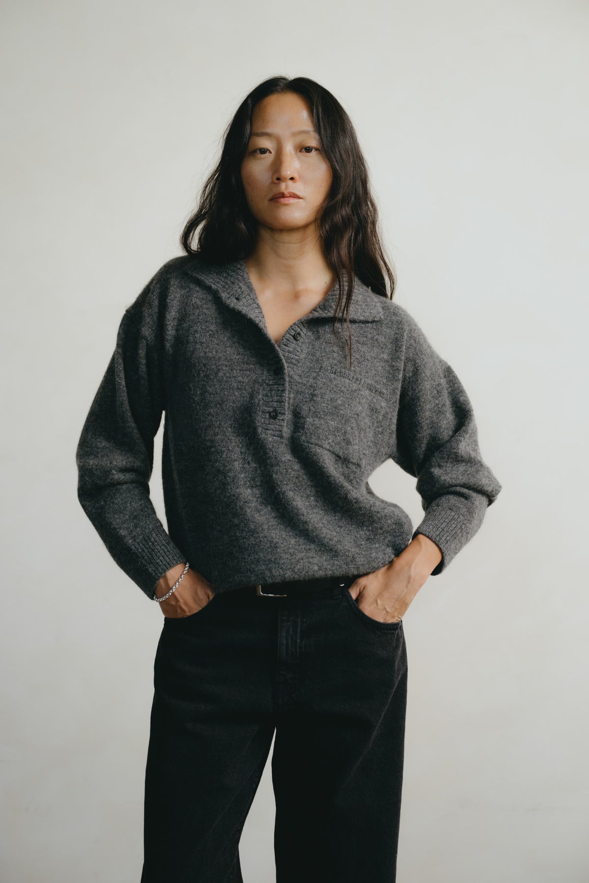 A woman wearing a dark gray knit polo sweater and black pants, standing against a plain light gray background.