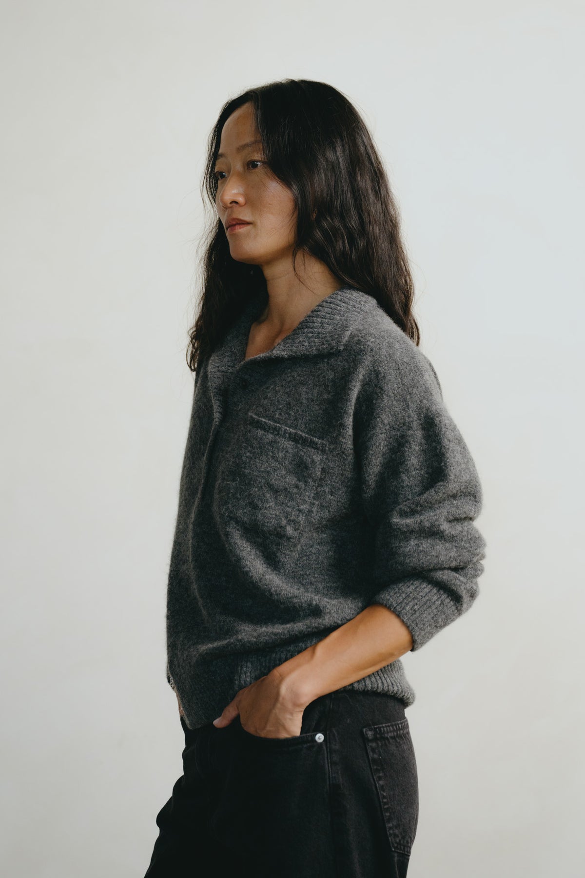 A woman with long dark hair is shown in profile wearing a dark gray textured knit sweater with a half-placket and a chest pocket, paired with black jeans, against a plain light gray background.