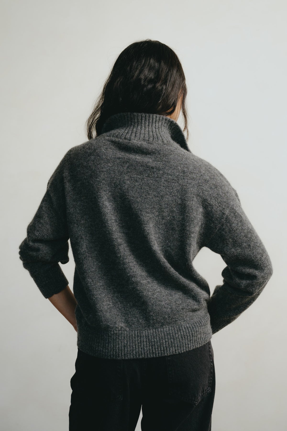 A person shown from behind wearing a dark gray ribbed turtleneck sweater and black pants against a plain light gray background.