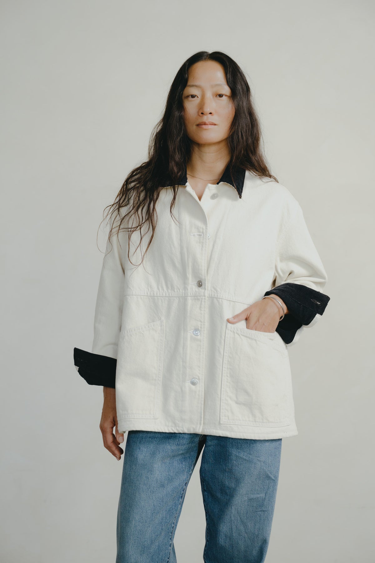 A woman modeling a white utility-style jacket with contrasting black collar and cuff details, paired with blue jeans, against a plain light gray studio background.