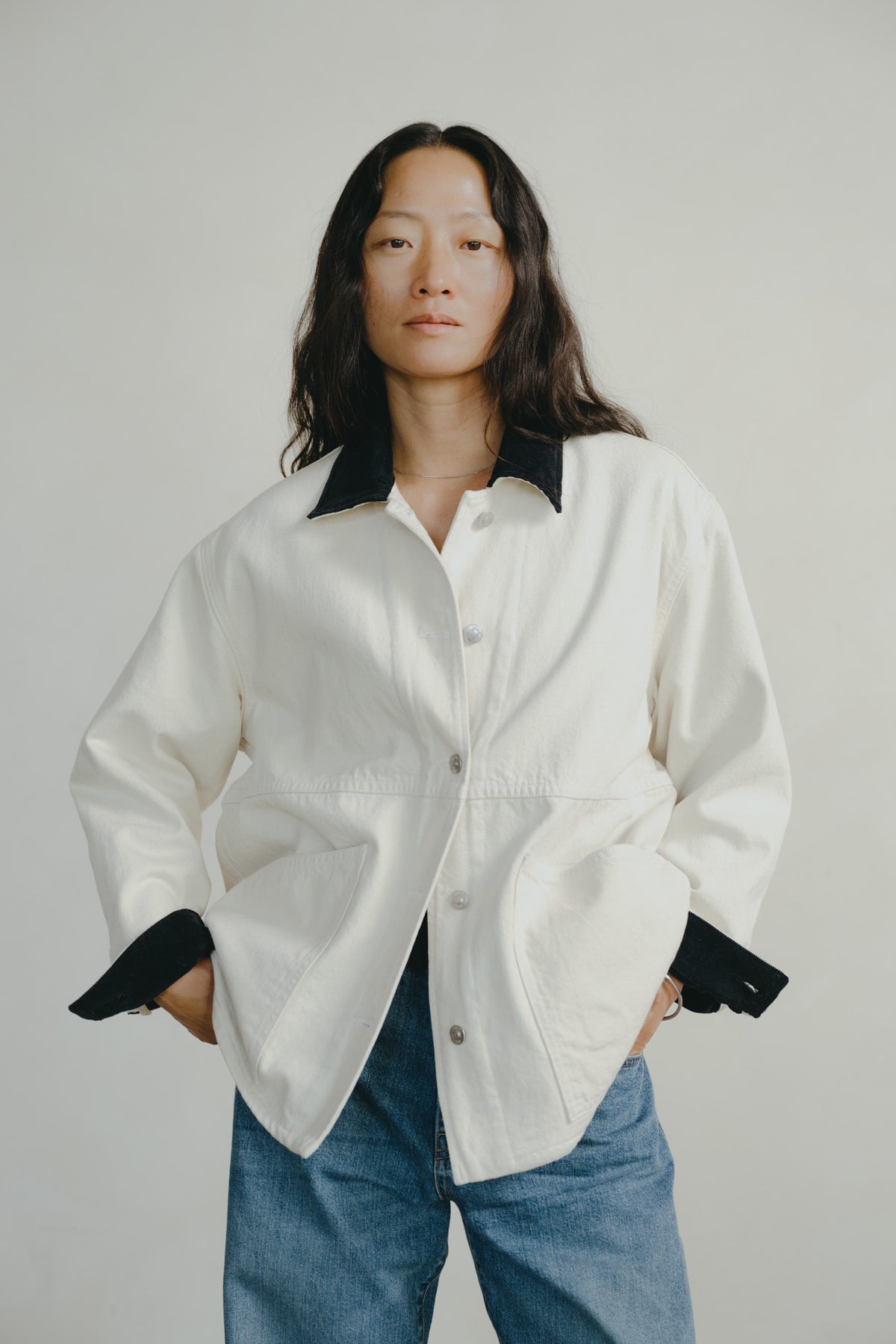 A model wearing a white button-up shirt jacket with black contrast collar, cuffs, and hem, paired with blue jeans, against a plain light gray background.