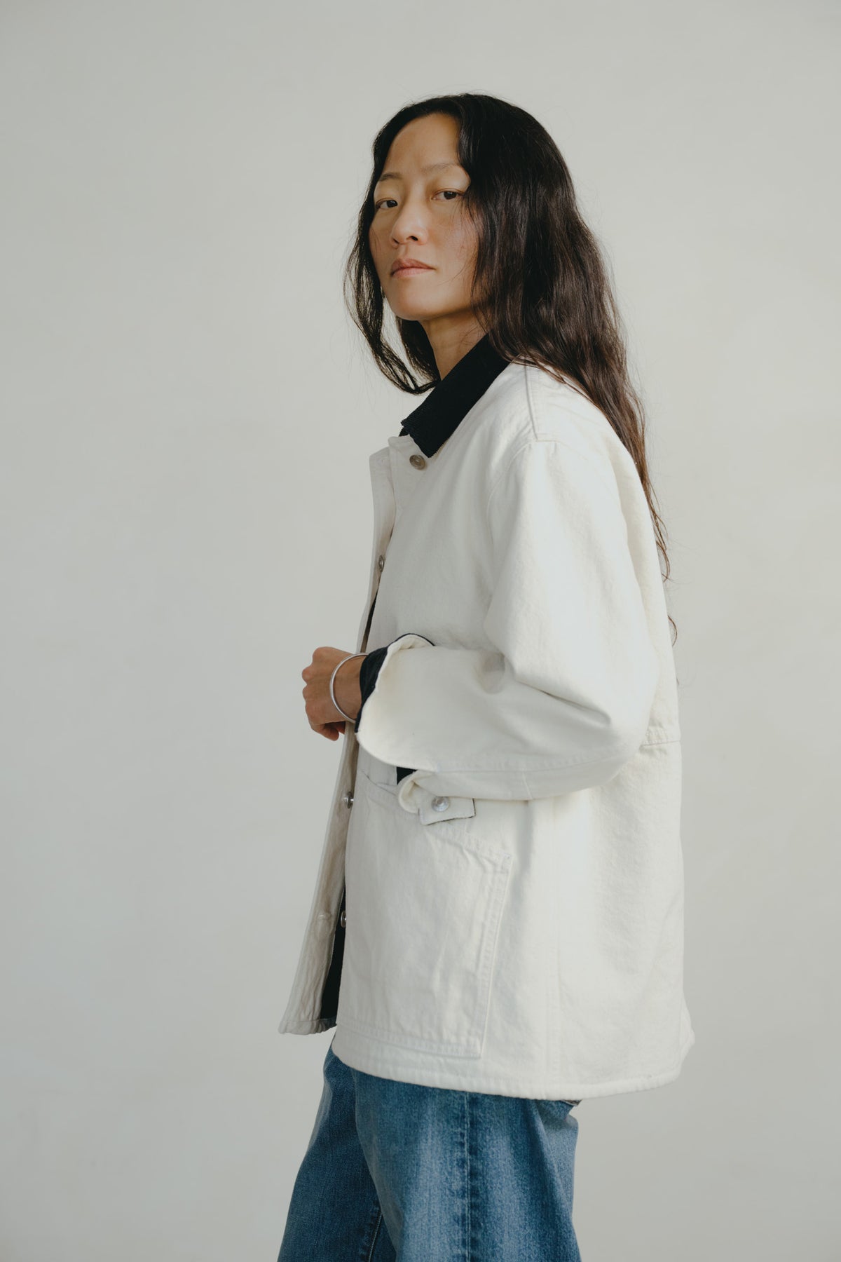 A model wearing an off-white canvas utility jacket with a black collar, paired with blue jeans, photographed against a neutral light gray background.