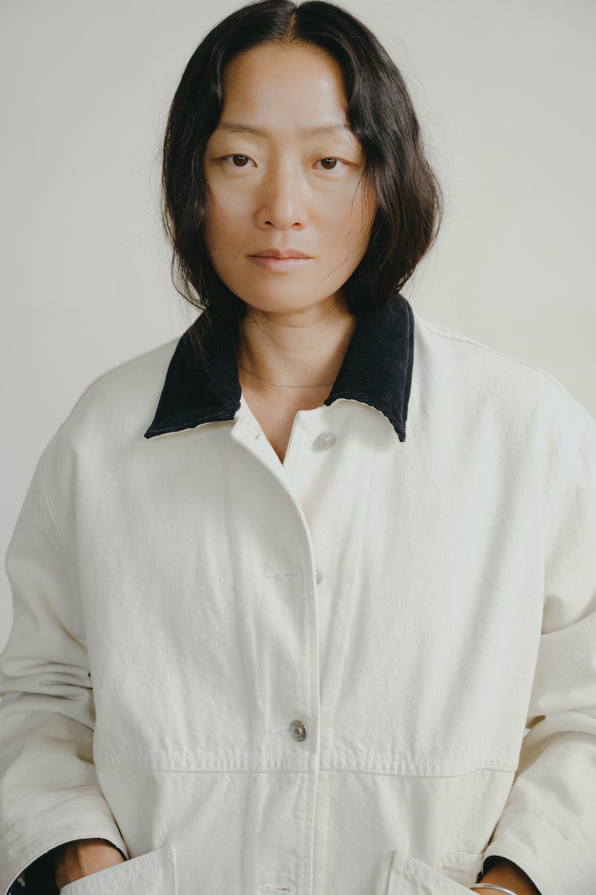 A woman with short dark hair wearing a white utility jacket with a contrasting black corduroy collar, photographed against a plain light gray background.