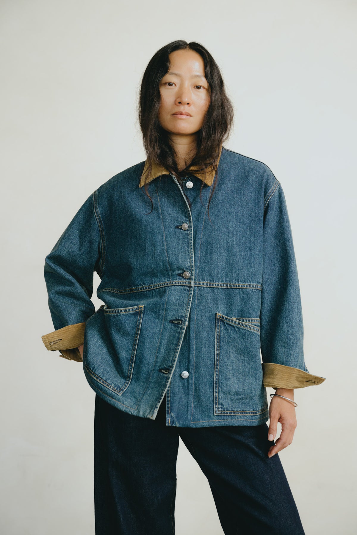 A model wearing a blue denim jacket with tan corduroy collar and cuffs, paired with dark blue pants, against a plain light gray background.
