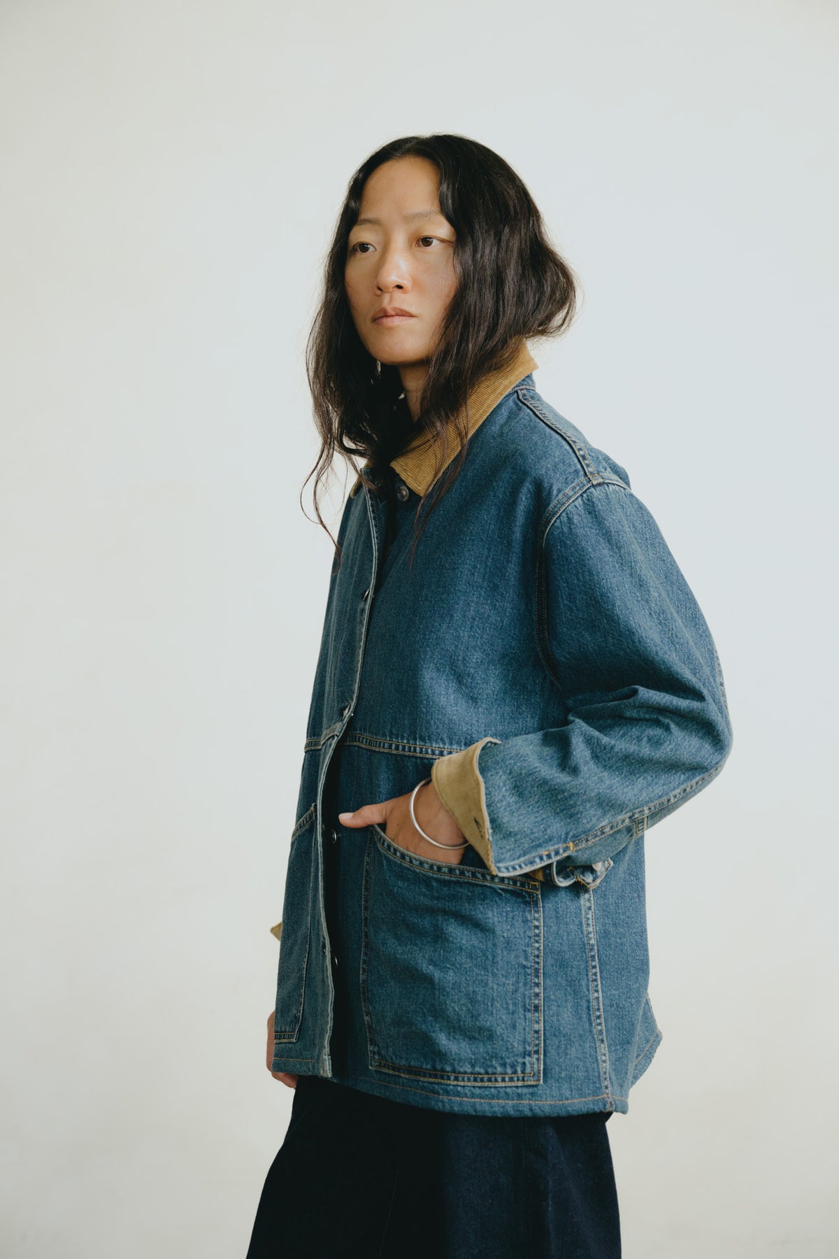 A model wearing a blue denim jacket with a tan collar and pocket lining, posed against a plain off-white studio background.