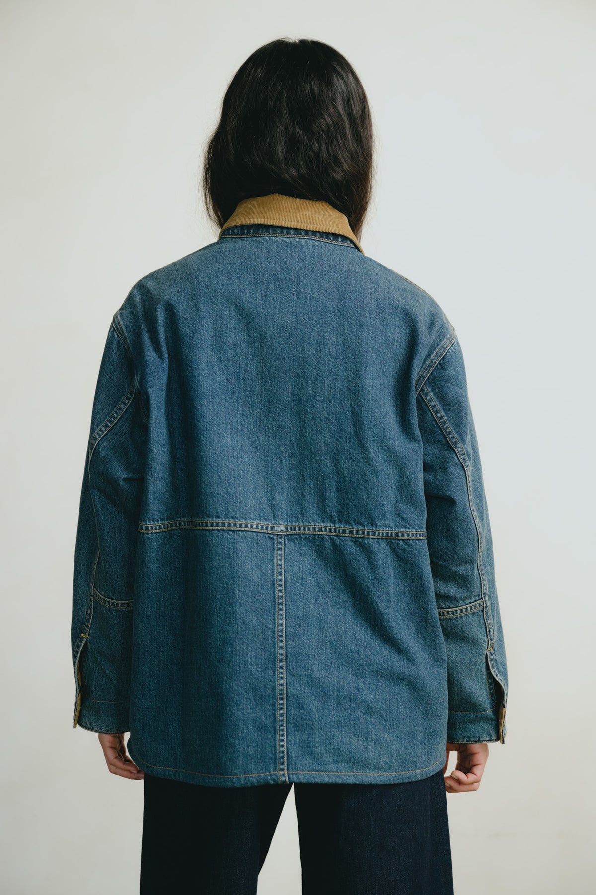 A person shown from the back wearing a blue denim jacket with a tan collar, paired with dark pants, against a plain light gray background.