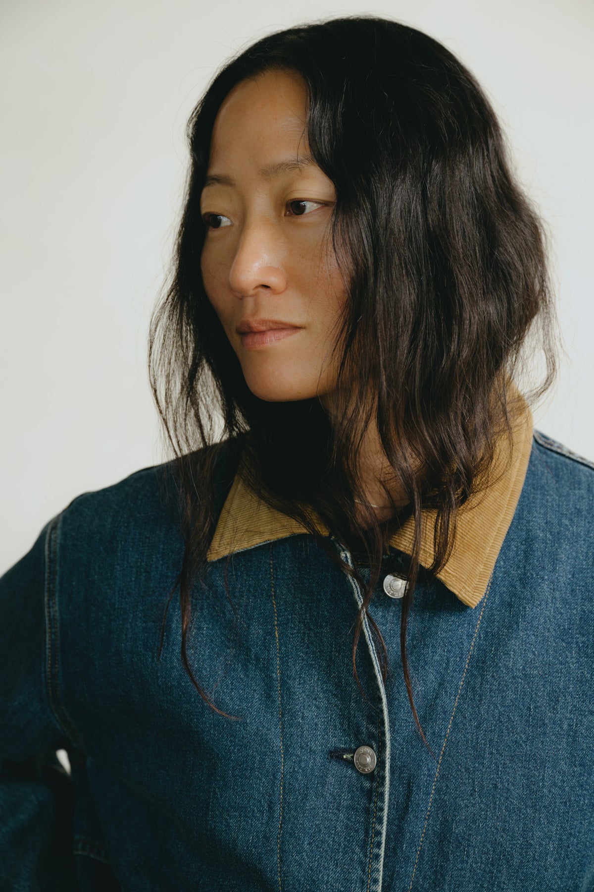 A close-up portrait of a woman with dark hair wearing a blue denim jacket with a tan collar, set against a plain light background.