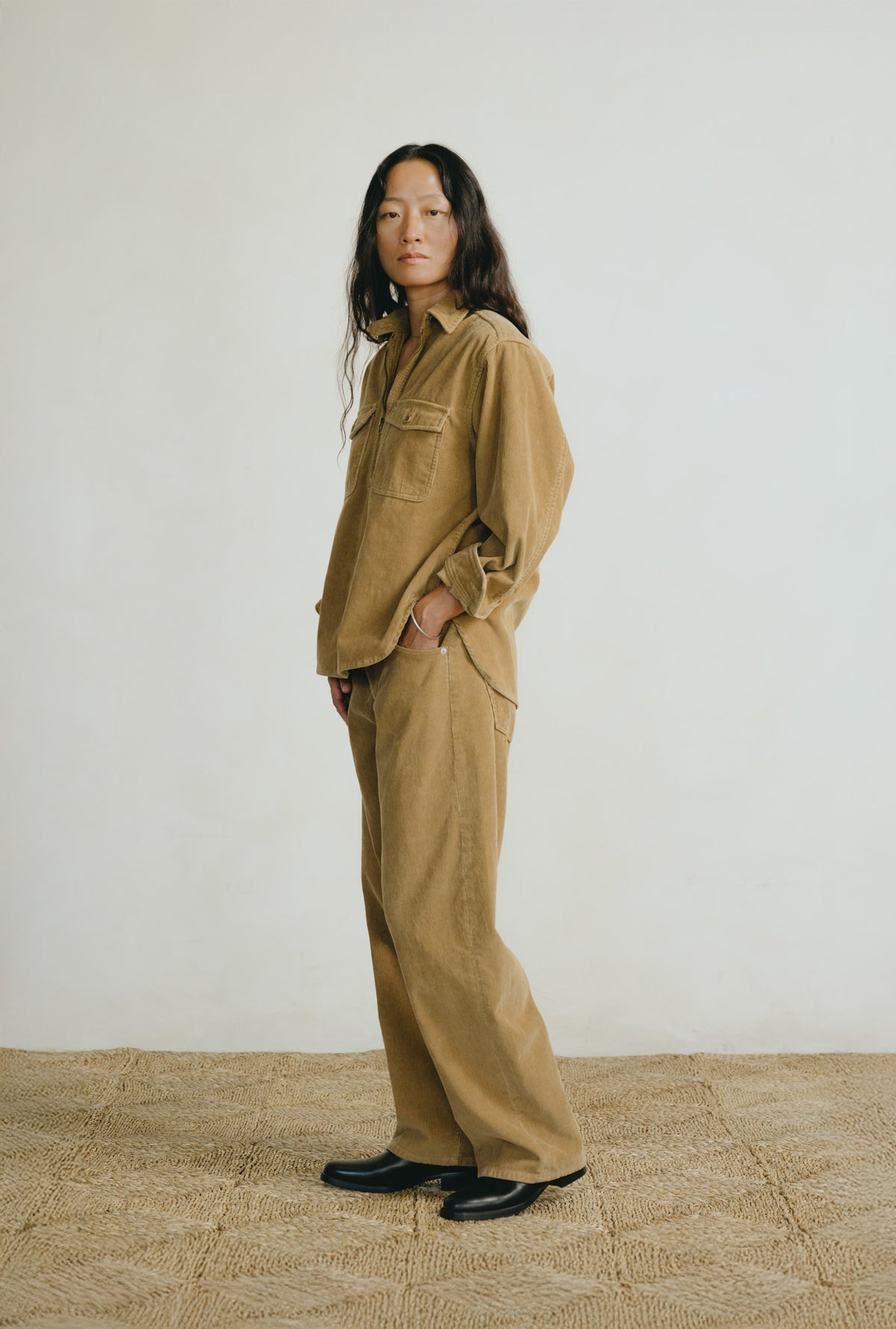 A model stands in profile wearing a matching tan corduroy utility jacket and wide-leg trousers set, paired with black shoes, against a plain off-white wall and textured beige floor.