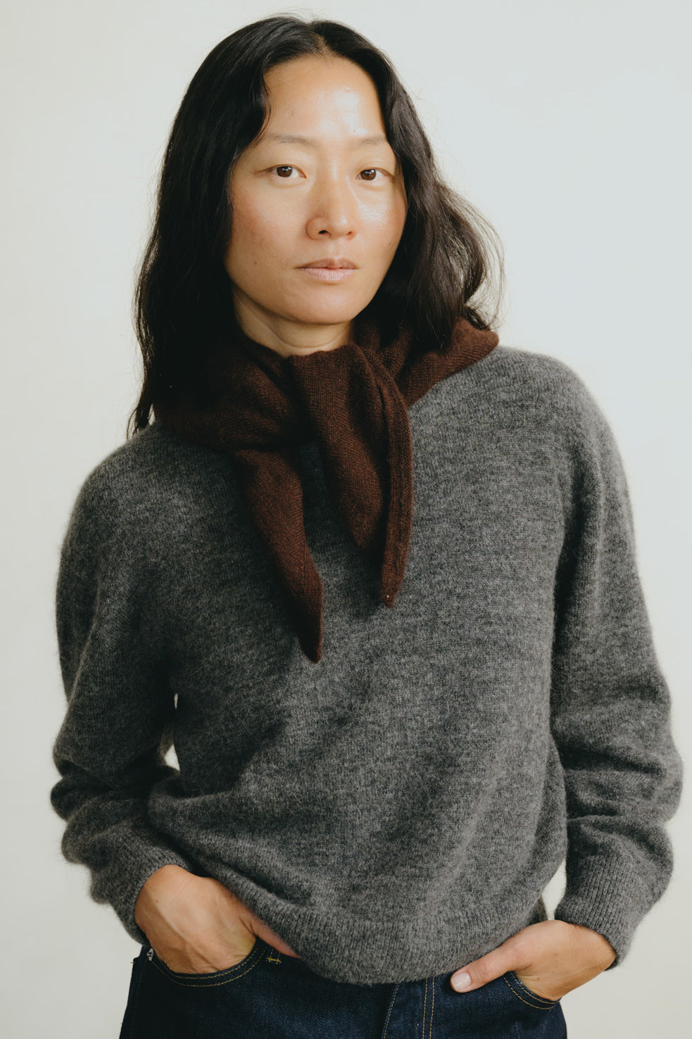 A woman wearing a dark gray knit sweater and a dark brown scarf, standing against a plain light gray background.