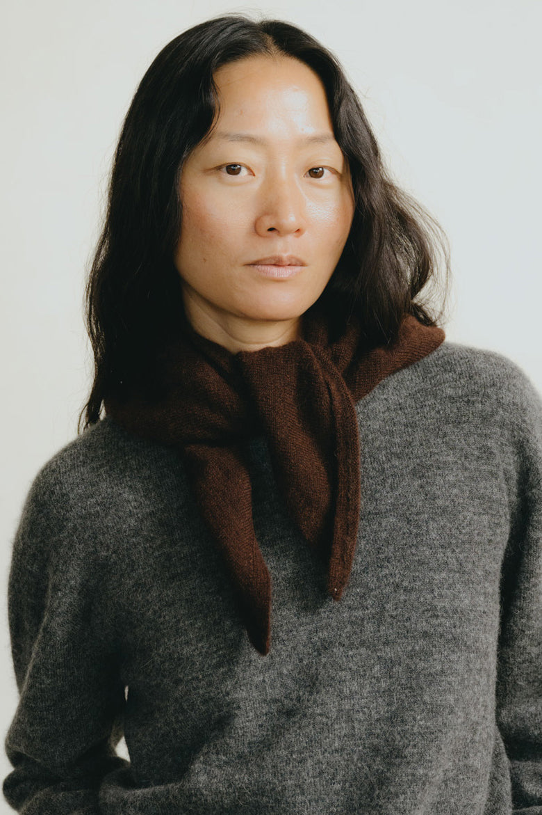 A portrait of a woman with long dark hair wearing a dark brown scarf and a gray knit sweater against a plain light background.