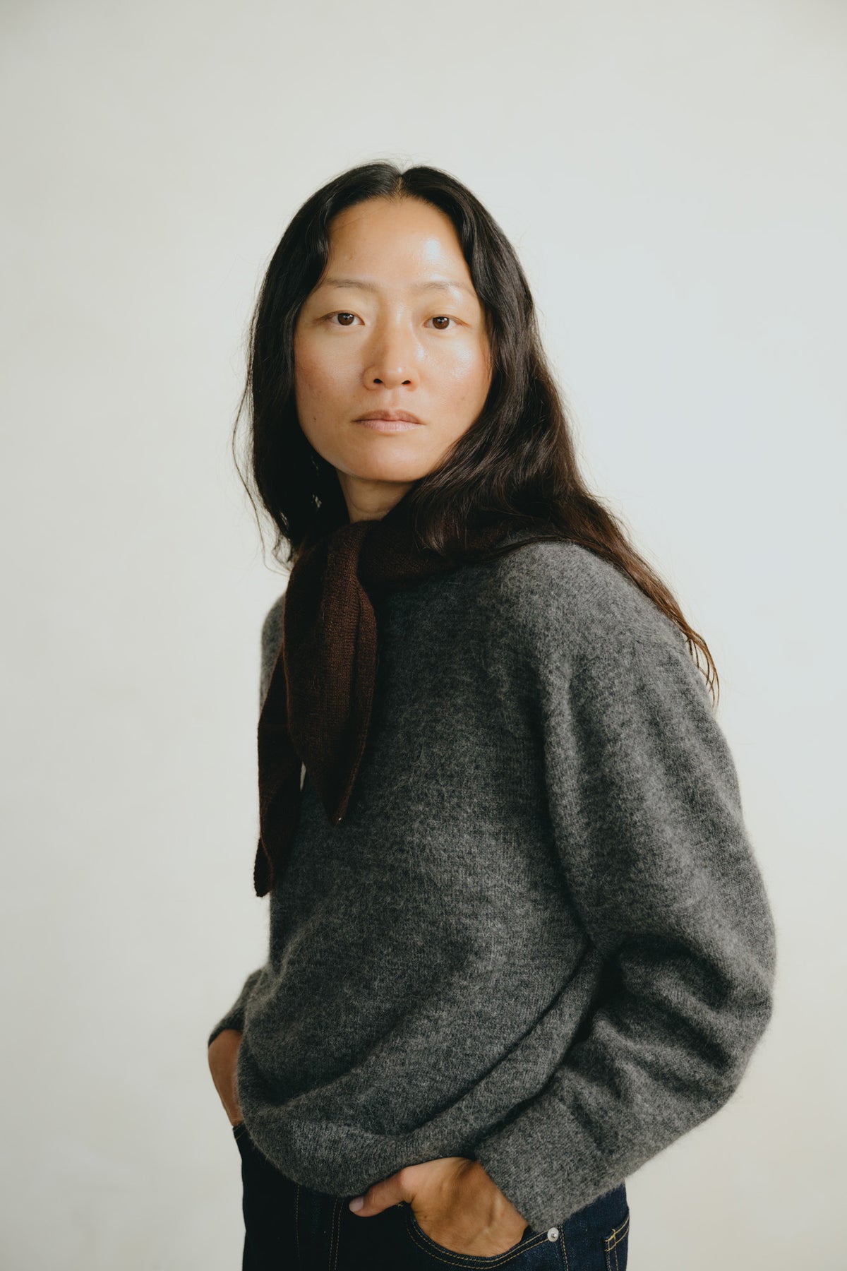 A woman with long dark hair wearing a textured gray knit sweater and a dark brown scarf, standing against a plain light gray background.