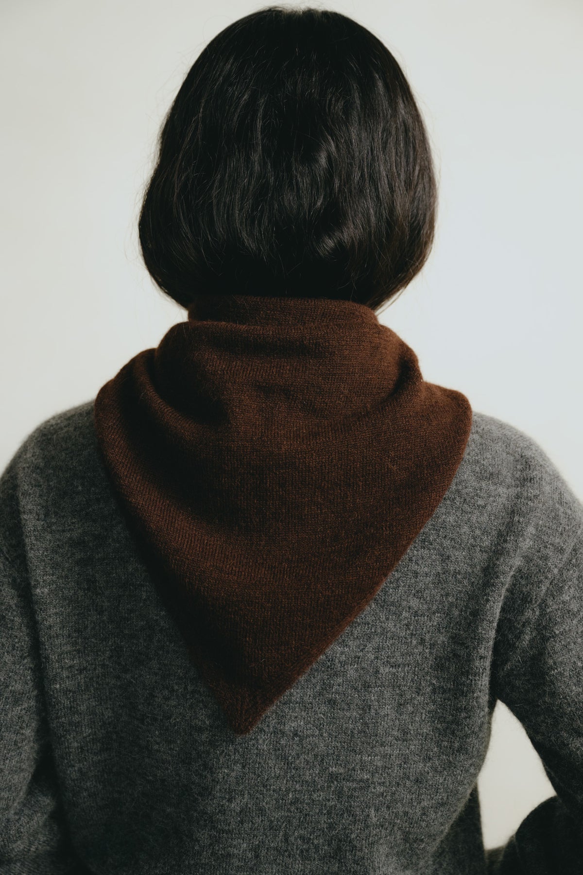 A person viewed from behind wearing a dark brown triangular knit scarf or neck warmer over a gray sweater, against a plain light background.