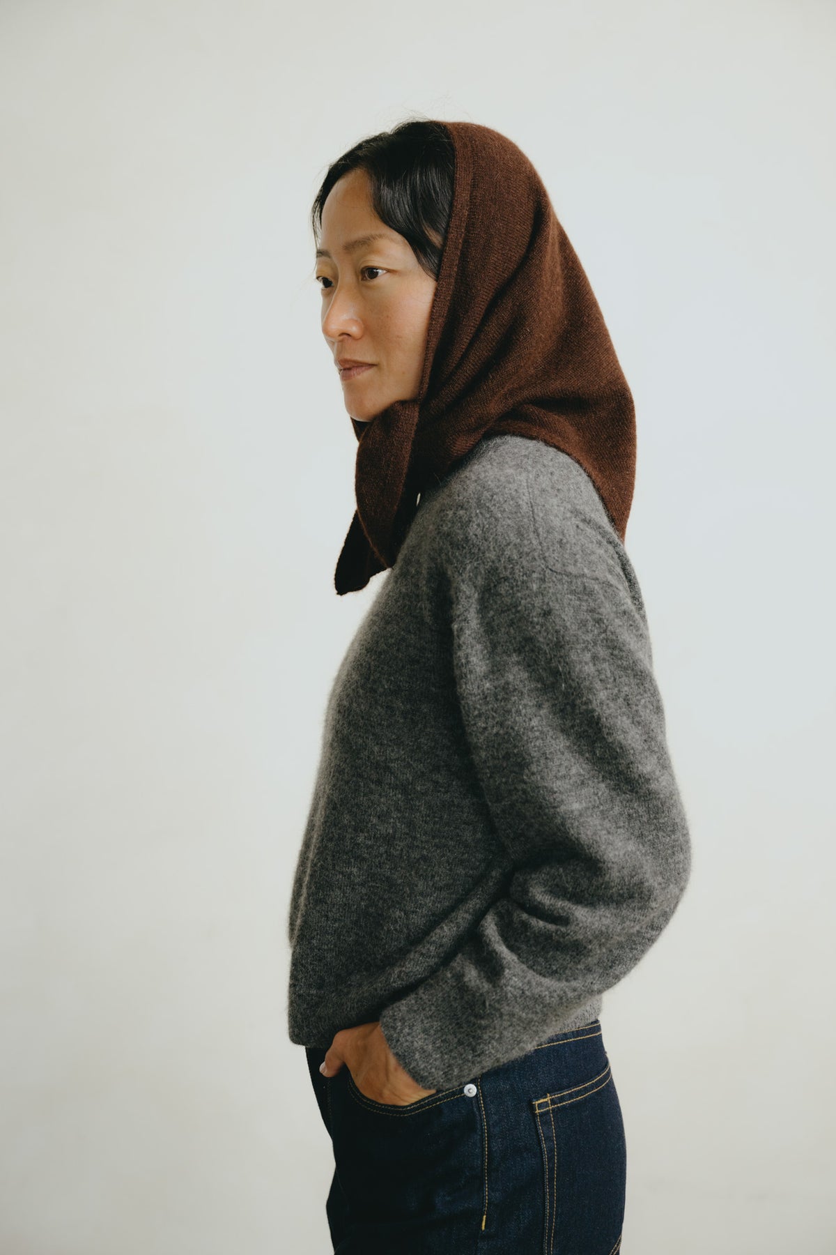 A woman shown in profile wearing a dark brown headscarf and a gray knit sweater, standing against a plain light gray background.