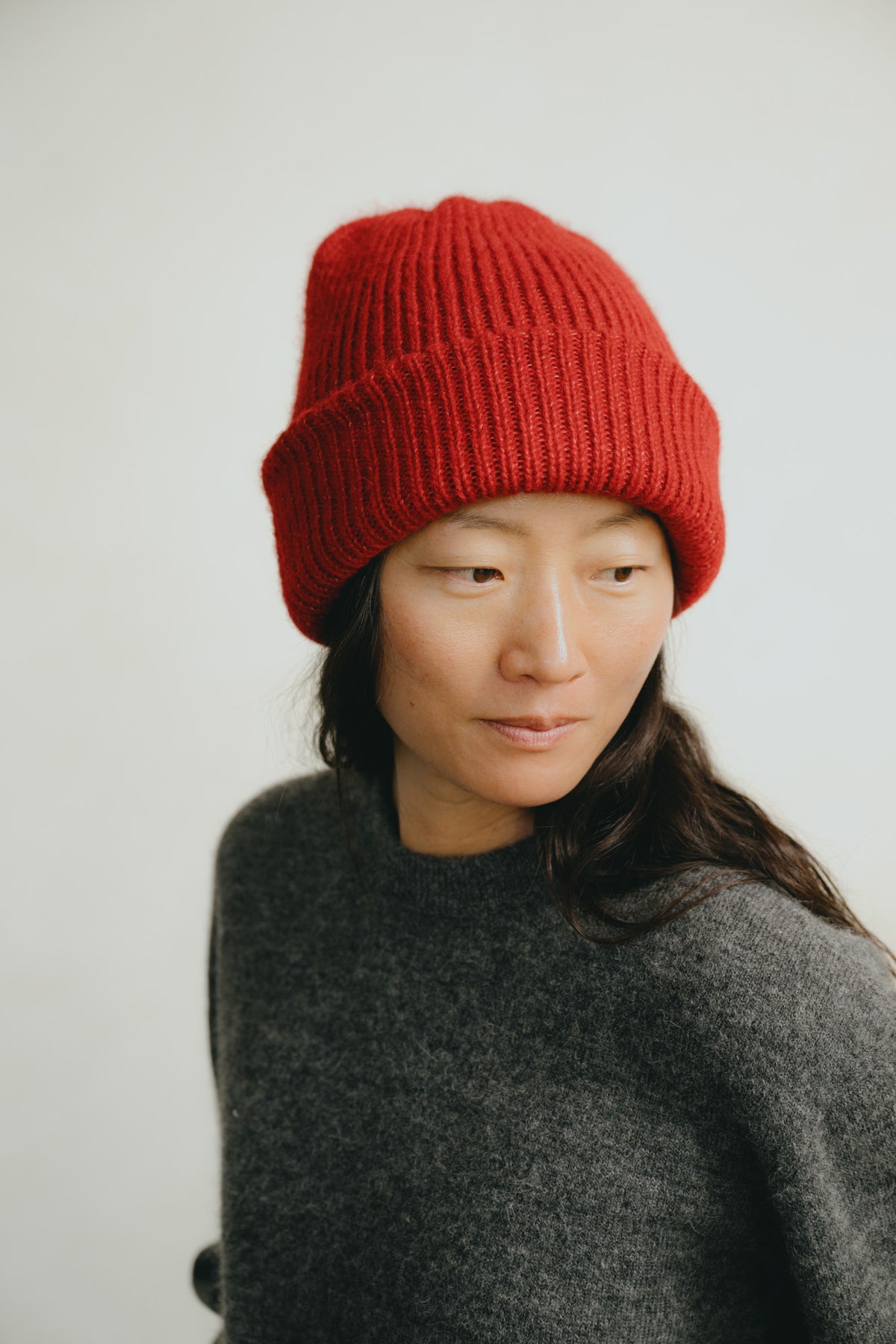 A woman wearing a bright red ribbed knit beanie and a dark gray knit sweater, photographed against a plain light gray background.